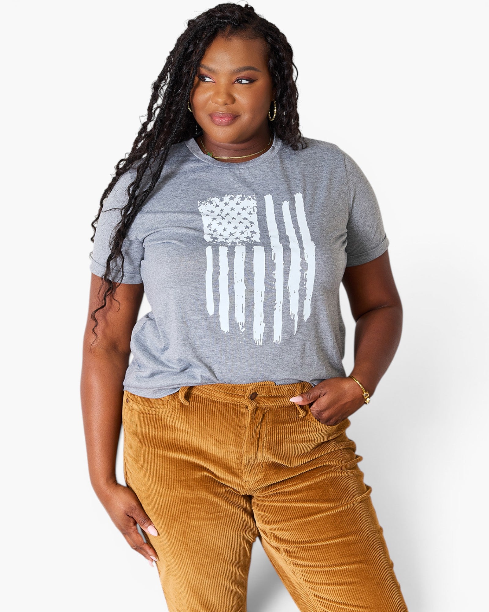 A woman modeling a heather gray short-sleeve t-shirt with a distressed white American flag graphic, paired with brown corduroy pants, against a plain white studio background.
