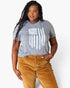 A woman modeling a heather gray short-sleeve t-shirt with a distressed white American flag graphic, paired with brown corduroy pants, against a plain white studio background.
