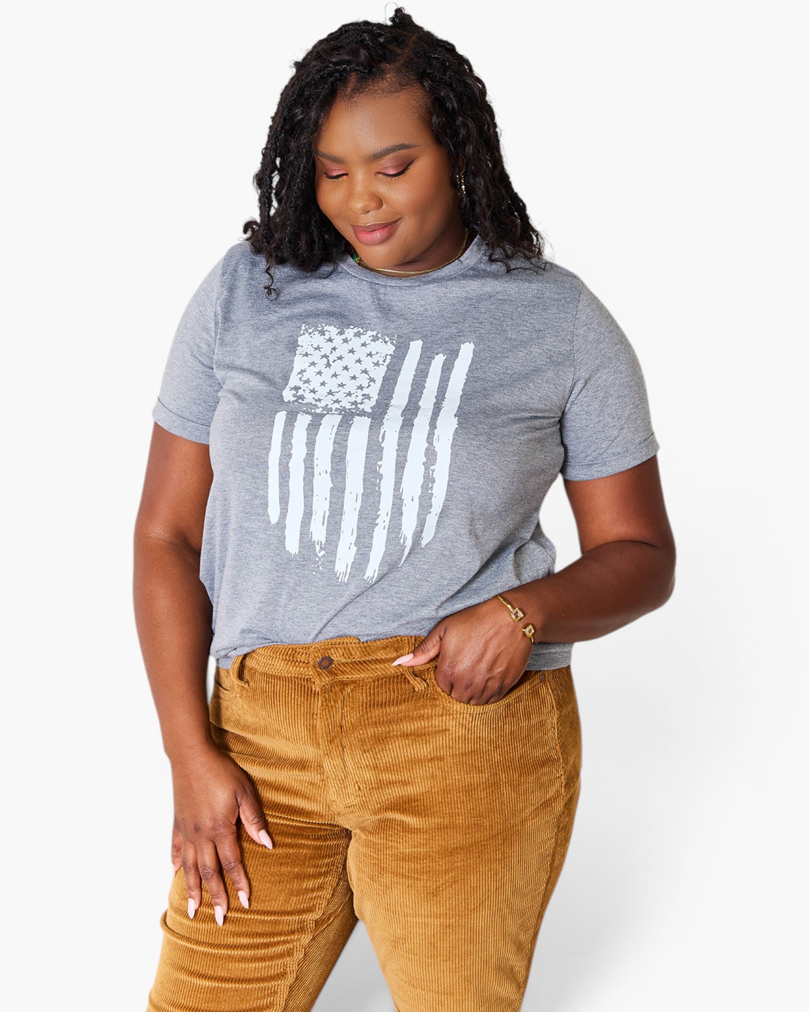 A woman wearing a heather gray t-shirt with a distressed white American flag graphic and mustard brown corduroy pants, photographed against a clean white background.