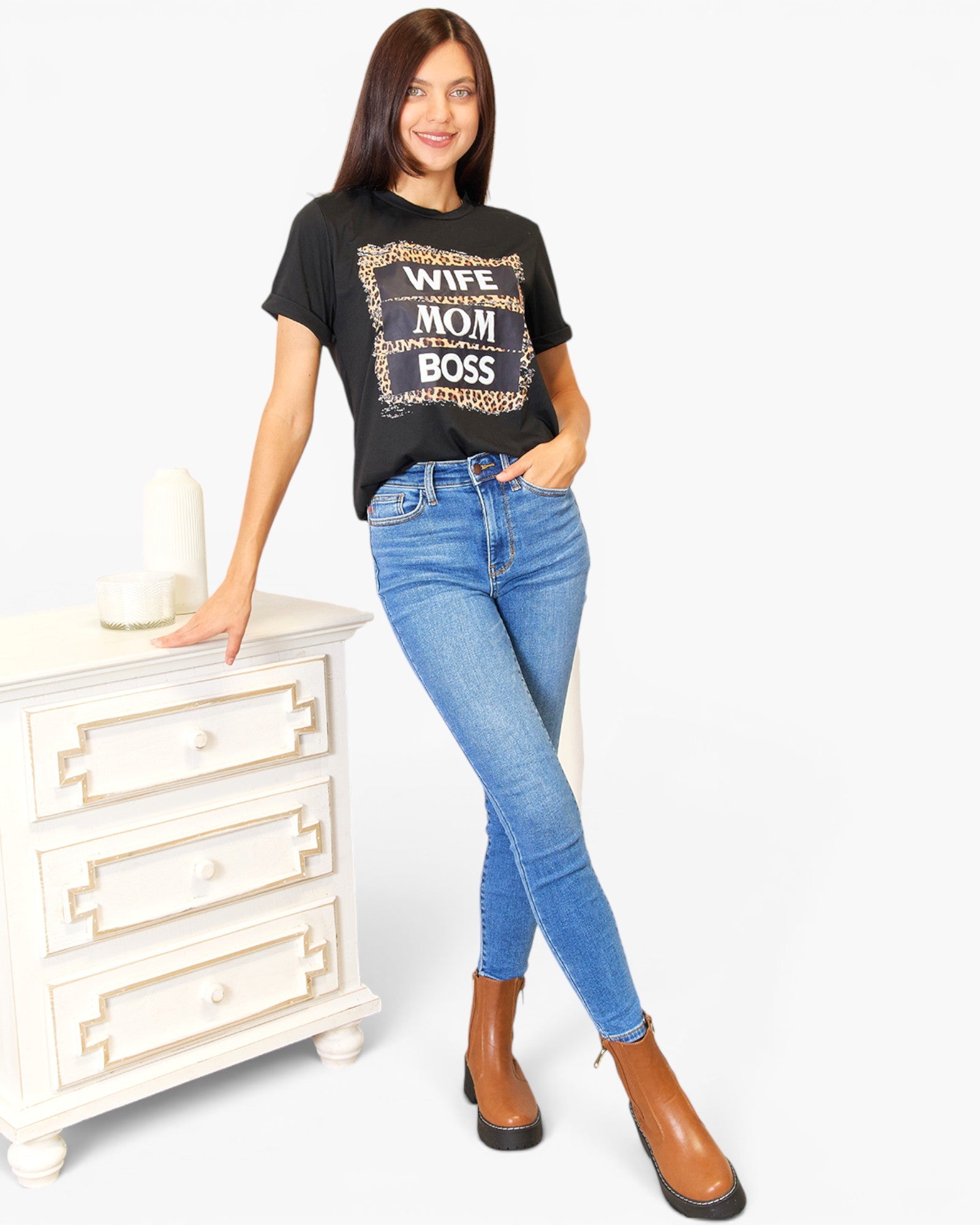 A woman modeling a black graphic t-shirt with a leopard print and white text that reads 'WIFE MOM BOSS', paired with blue skinny jeans and brown ankle boots, standing next to a white three-drawer cabinet.