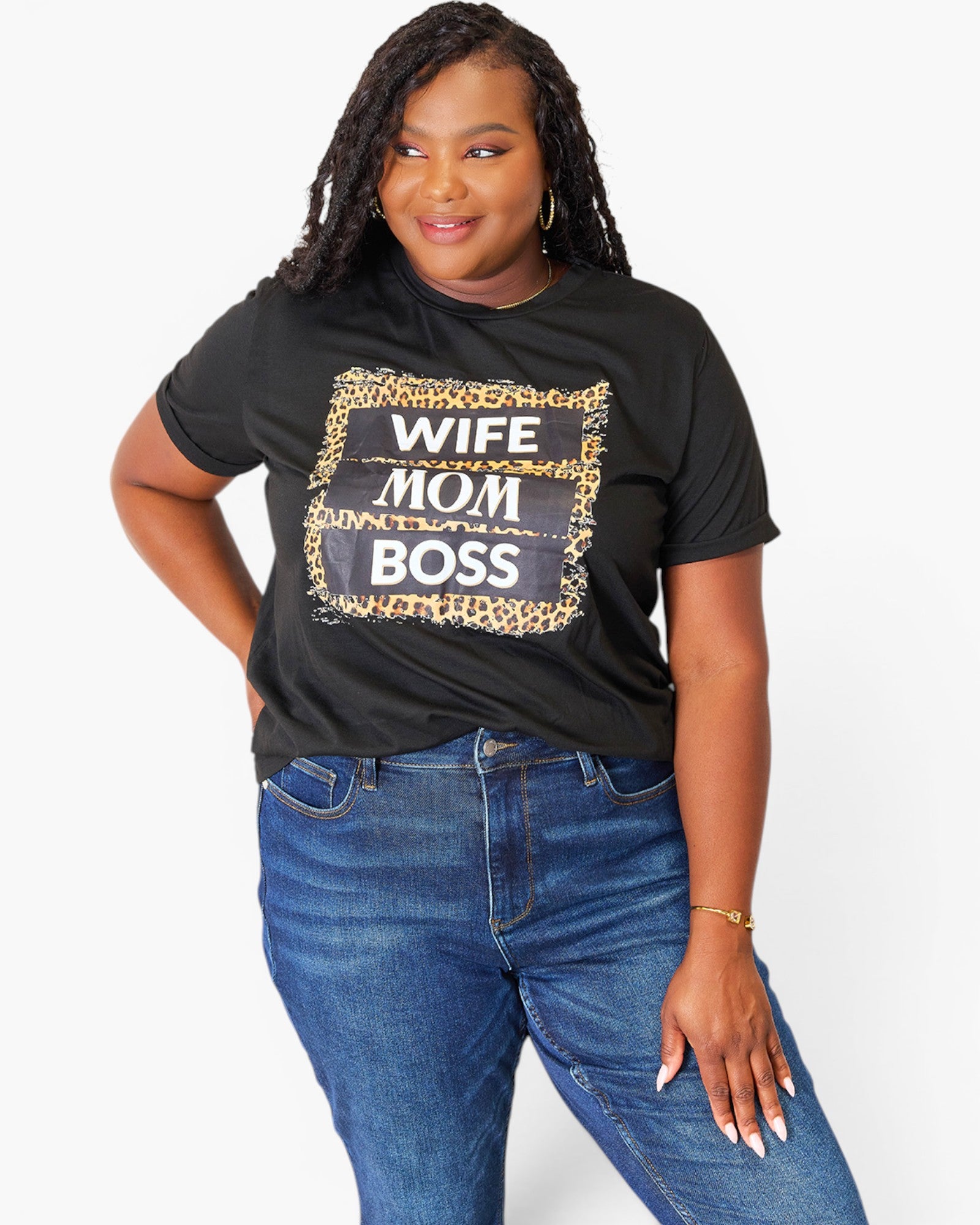 A woman modeling a black graphic t-shirt with a leopard print and metallic text that reads 'WIFE MOM BOSS', paired with blue jeans against a plain white background.