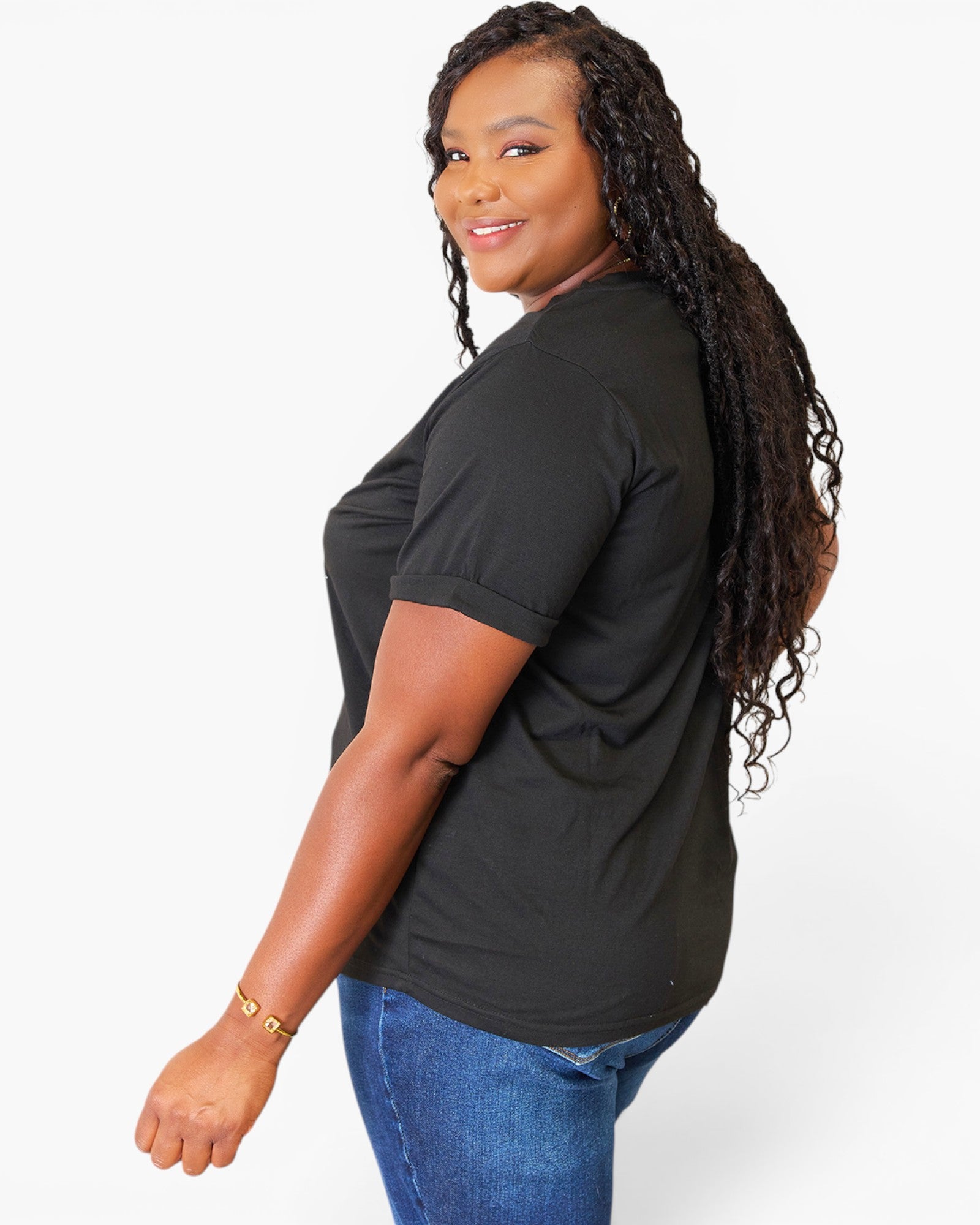 A woman with long, curly black hair is shown from a side-back angle wearing a black short-sleeve t-shirt and blue jeans against a plain white studio background.