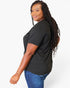 A woman with long, curly black hair is shown from a side-back angle wearing a black short-sleeve t-shirt and blue jeans against a plain white studio background.