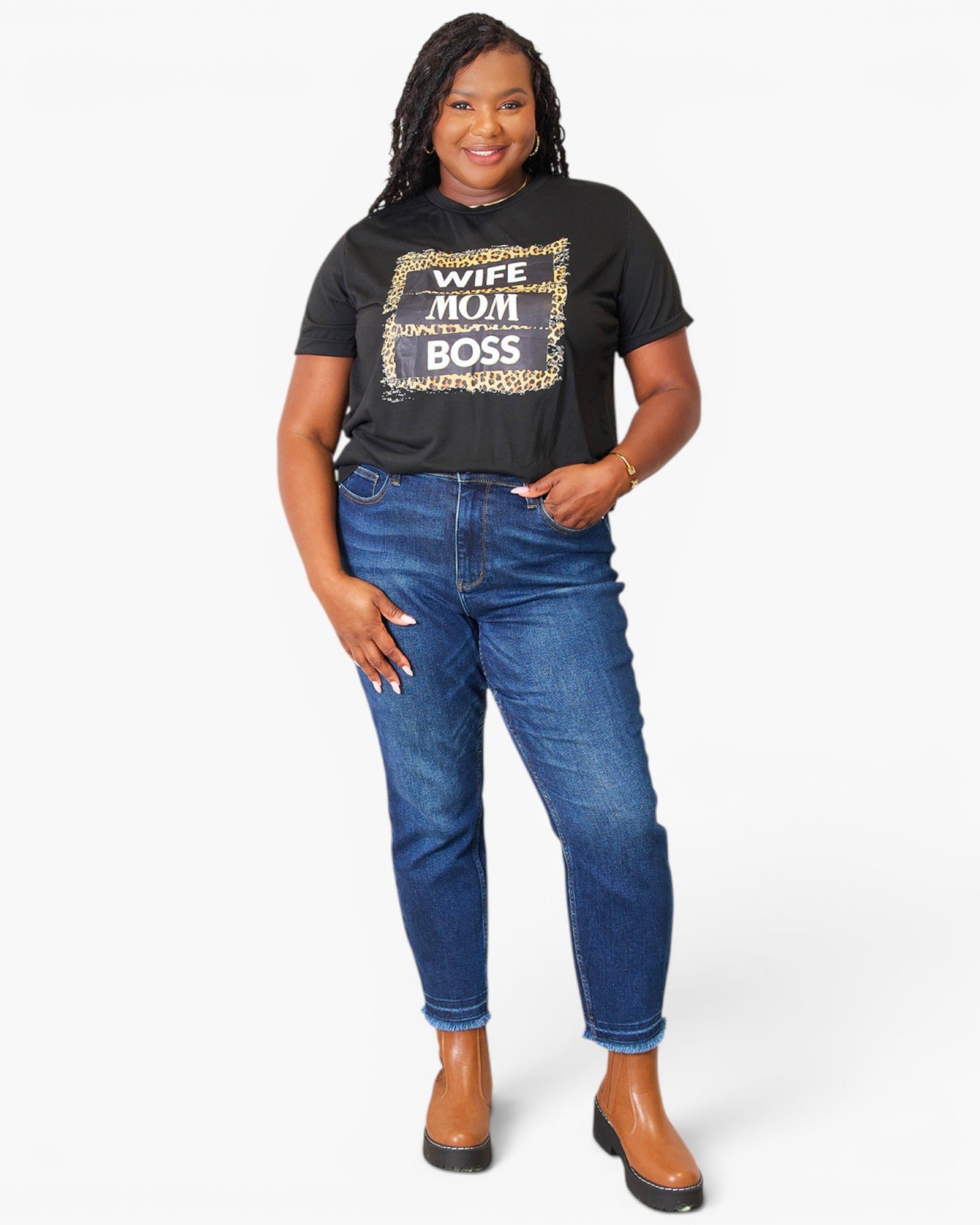 A woman modeling a black graphic t-shirt with a leopard print border and the words 'WIFE MOM BOSS' in white and gold, paired with blue jeans and brown ankle boots, against a light gray background.