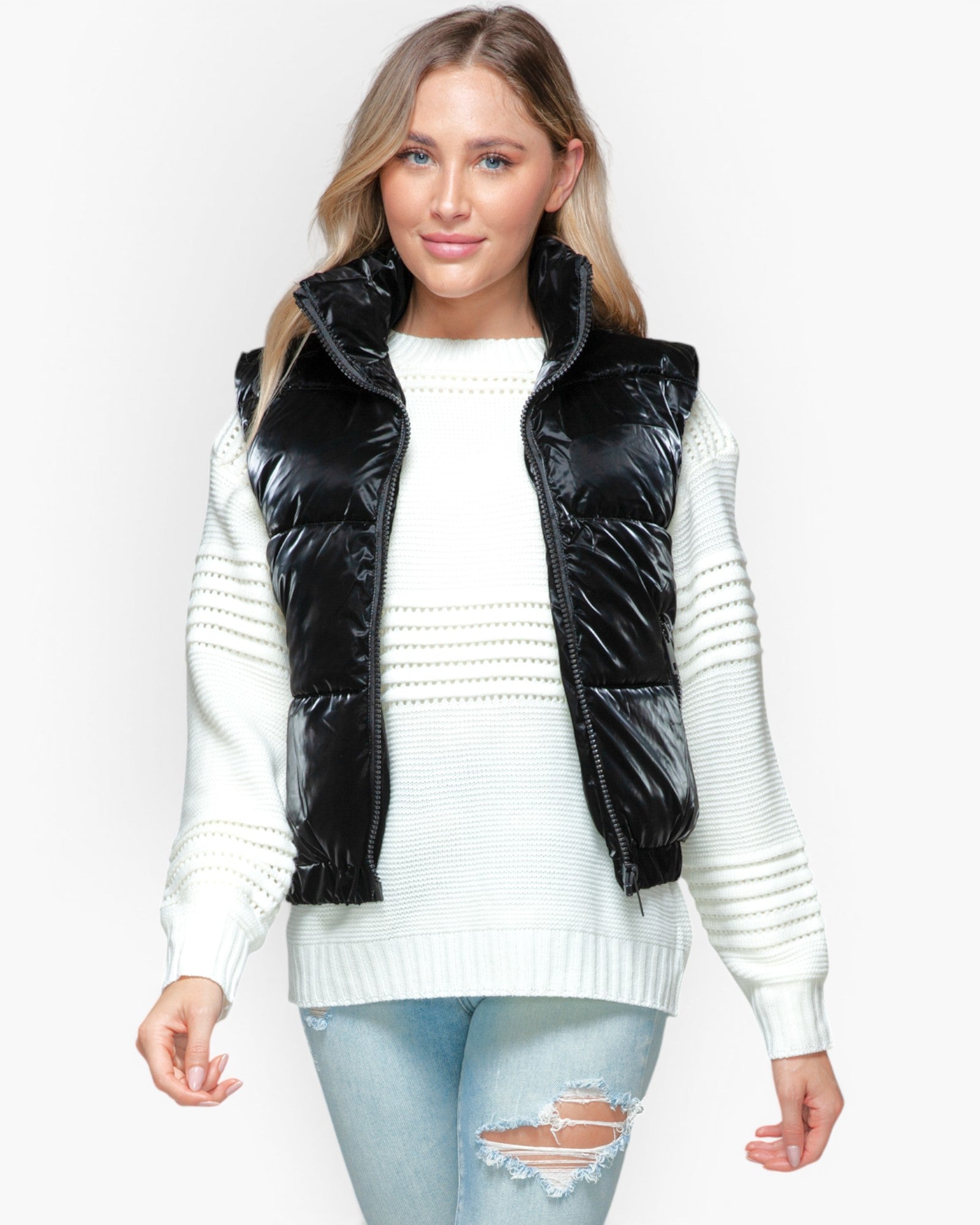 A woman modeling a black puffer vest over a white knit sweater and light blue ripped jeans, photographed against a plain light gray studio background.