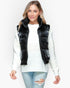 A woman modeling a black puffer vest over a white knit sweater and light blue ripped jeans, photographed against a plain light gray studio background.