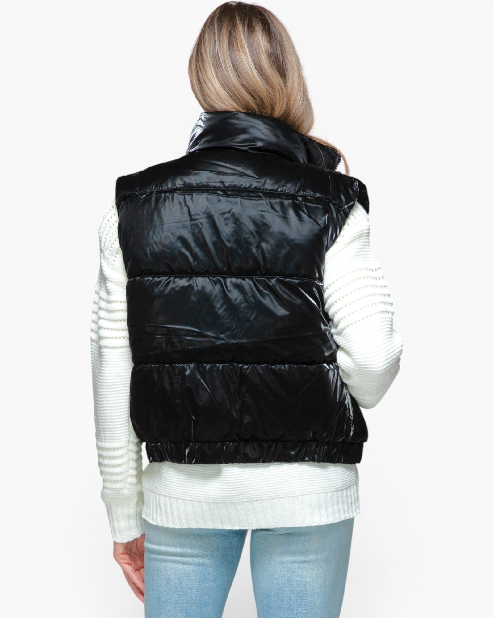 A woman shown from the back wearing a shiny black puffer vest over a white ribbed sweater and light blue jeans, against a plain white studio background.