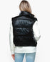 A woman shown from the back wearing a shiny black puffer vest over a white ribbed sweater and light blue jeans, against a plain white studio background.