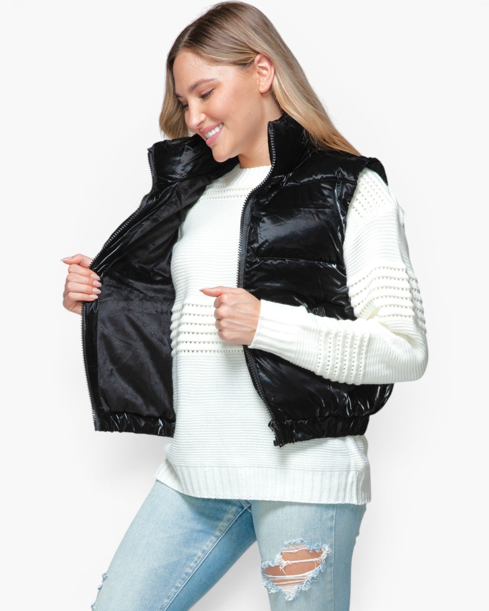 A woman wearing a shiny black quilted puffer vest over a white ribbed knit sweater and light blue distressed jeans, shown against a plain light gray studio background.