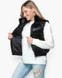 A woman wearing a shiny black quilted puffer vest over a white ribbed knit sweater and light blue distressed jeans, shown against a plain light gray studio background.