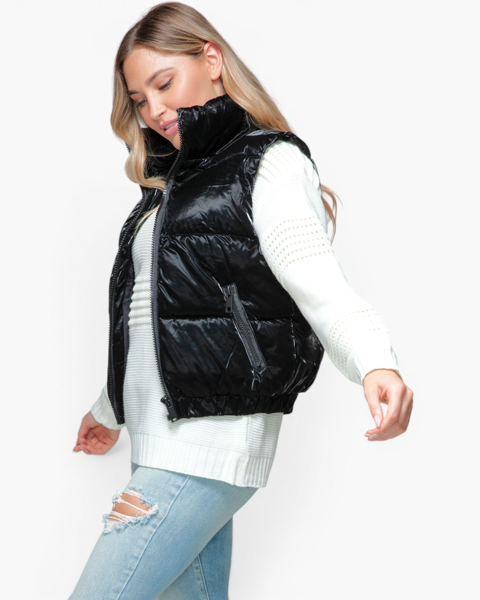 A woman wearing a shiny black quilted puffer vest over a white knit sweater and light blue ripped jeans, photographed against a plain light gray studio background.