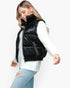 A woman wearing a shiny black quilted puffer vest over a white knit sweater and light blue ripped jeans, photographed against a plain light gray studio background.