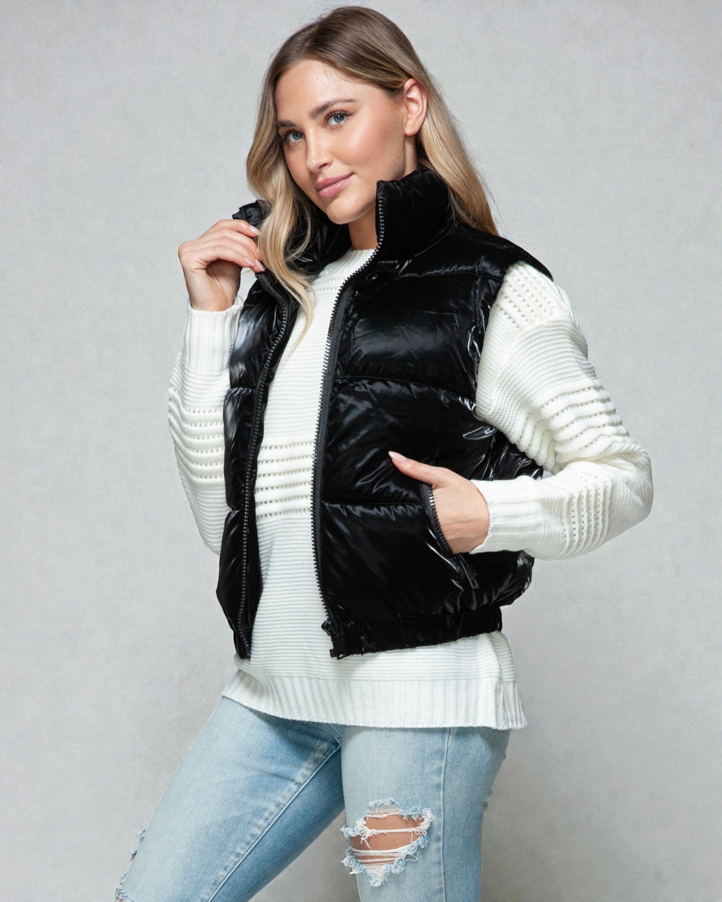 Woman wearing a black puffer vest over a white sweater and light blue jeans on a gray background