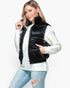 A woman modeling a black quilted puffer vest over a white ribbed knit sweater and light blue distressed jeans, photographed against a light gray studio background.