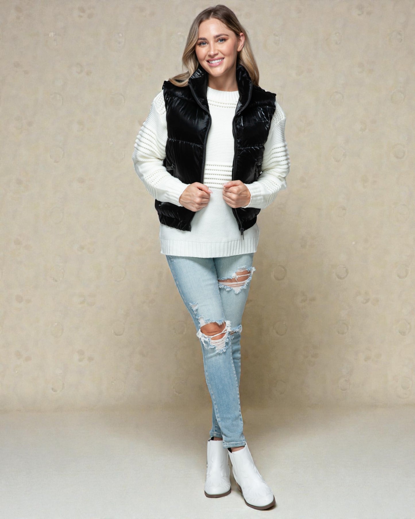 Woman wearing a black puffer vest over a white sweater, light blue jeans, and white boots on a beige background.