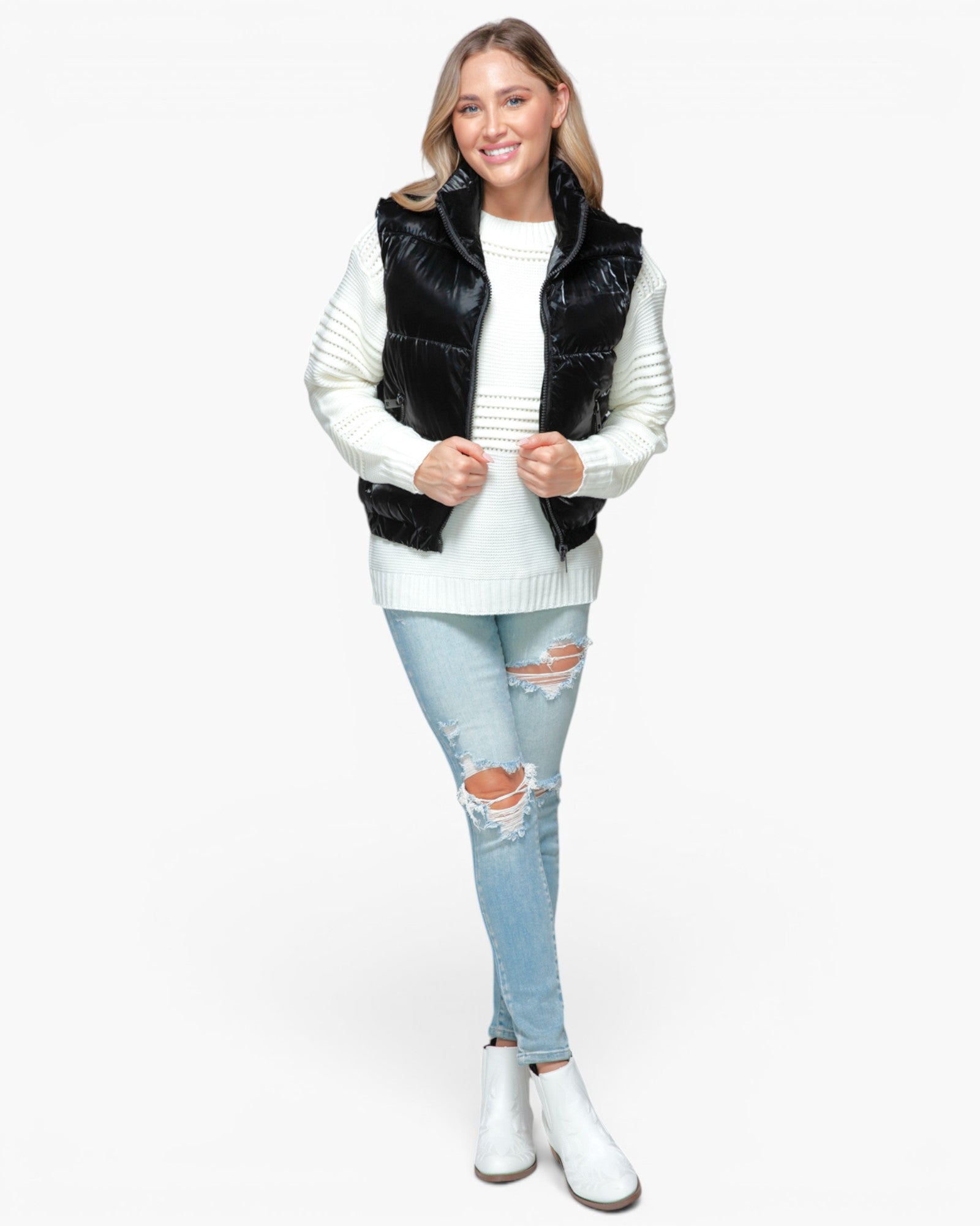 A woman modeling a black puffer vest over a white ribbed sweater, paired with light blue distressed jeans and white ankle boots, against a plain white background.