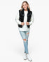 A woman modeling a black puffer vest over a white ribbed sweater, paired with light blue distressed jeans and white ankle boots, against a plain white background.