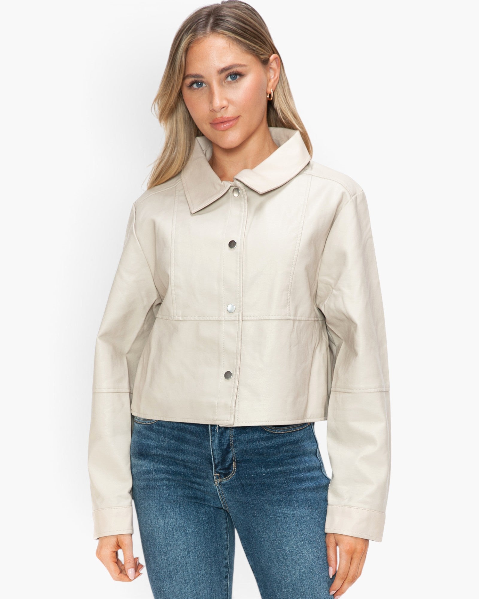 A woman modeling a cropped, light beige faux-leather jacket with a pointed collar and button-front closure, paired with blue jeans against a plain white studio background.
