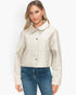 A woman modeling a cropped, light beige faux-leather jacket with a pointed collar and button-front closure, paired with blue jeans against a plain white studio background.