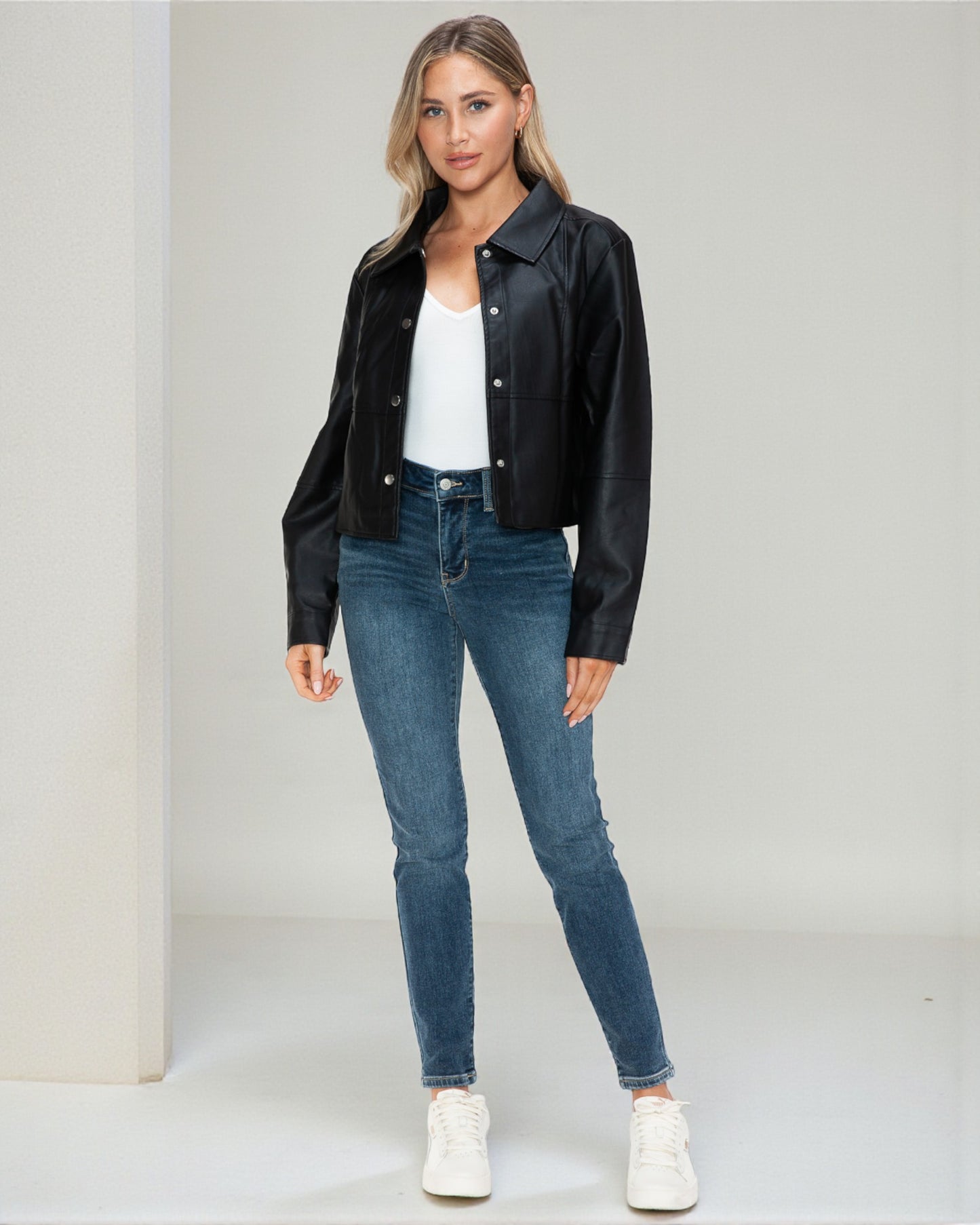 Woman wearing a black leather jacket, white shirt, blue jeans, and white sneakers on a plain background