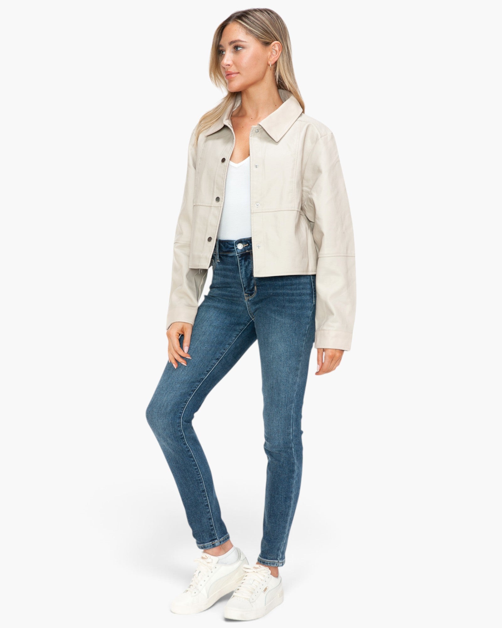 A woman modeling a light beige cropped jacket over a white top and blue skinny jeans, paired with white sneakers, against a plain white studio background.