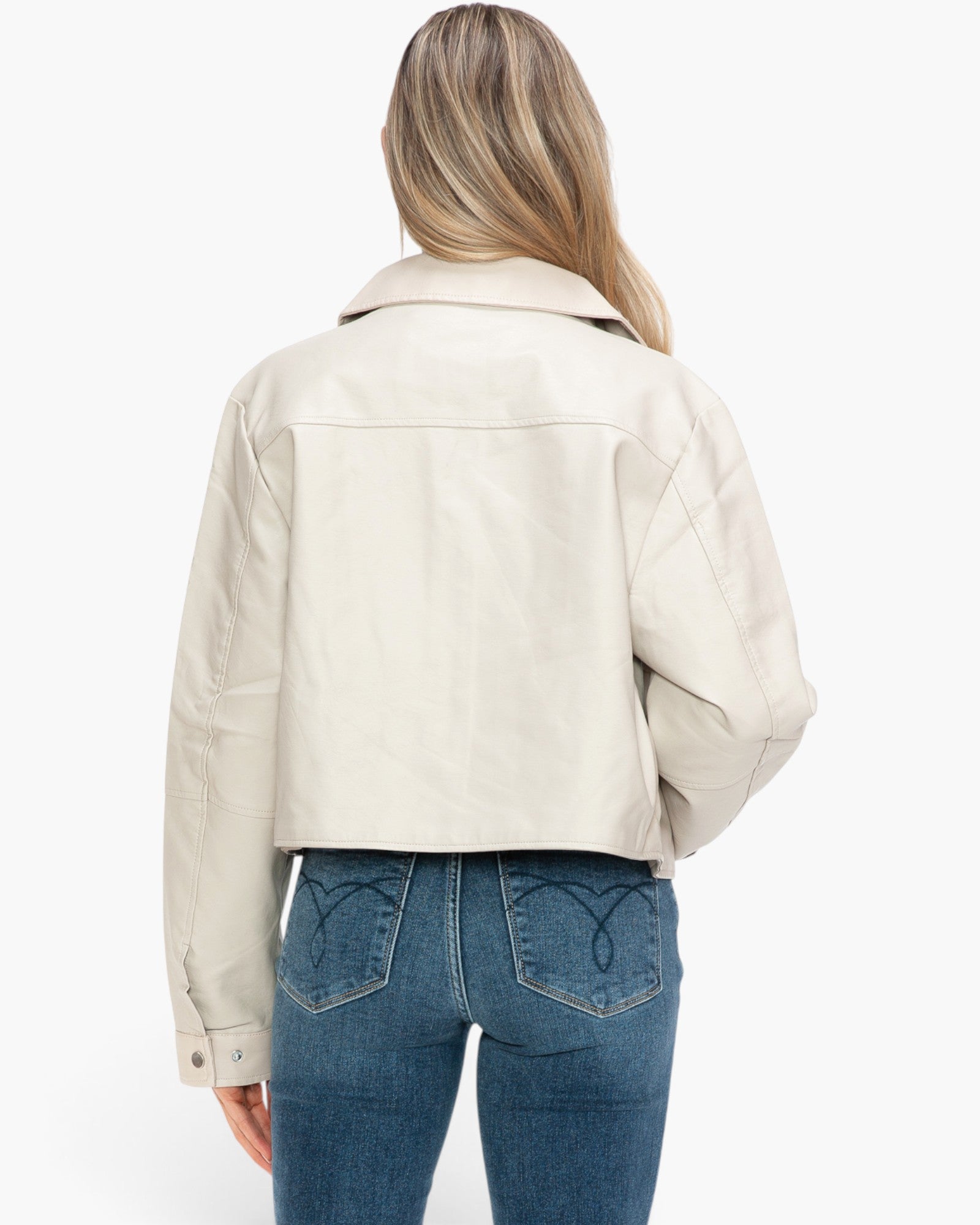 A woman viewed from the back wearing a cropped off-white leather jacket and blue jeans against a plain white background.