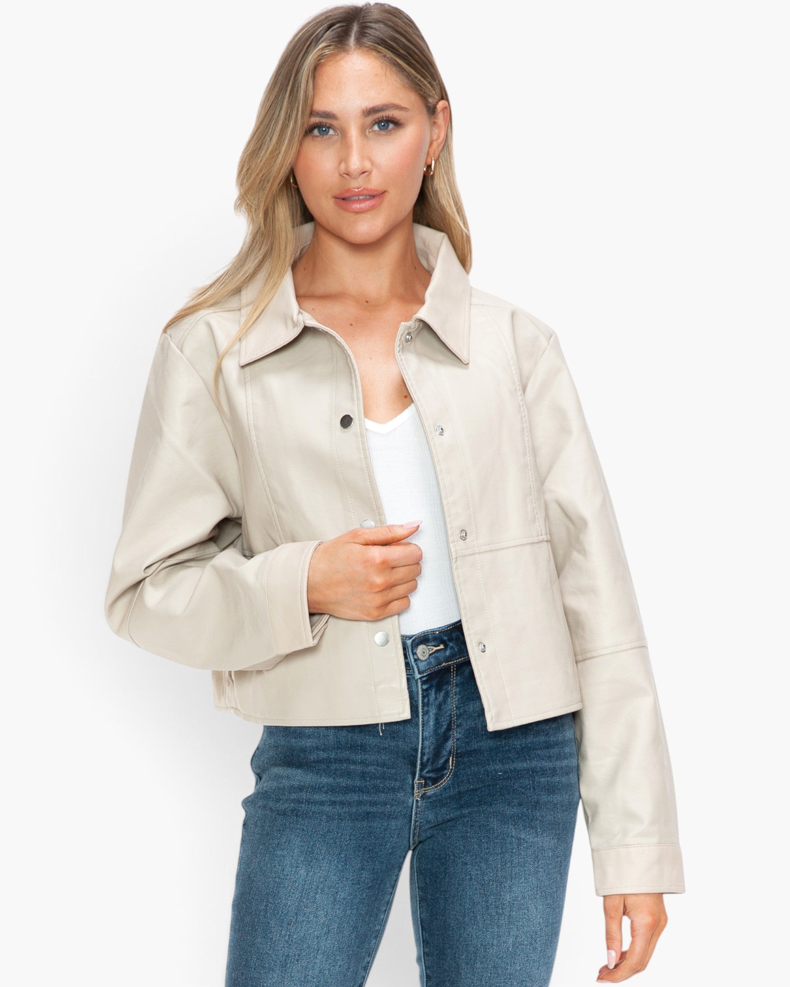 A woman modeling a cropped, light beige faux-leather jacket over a white top and blue jeans, set against a plain white studio background.