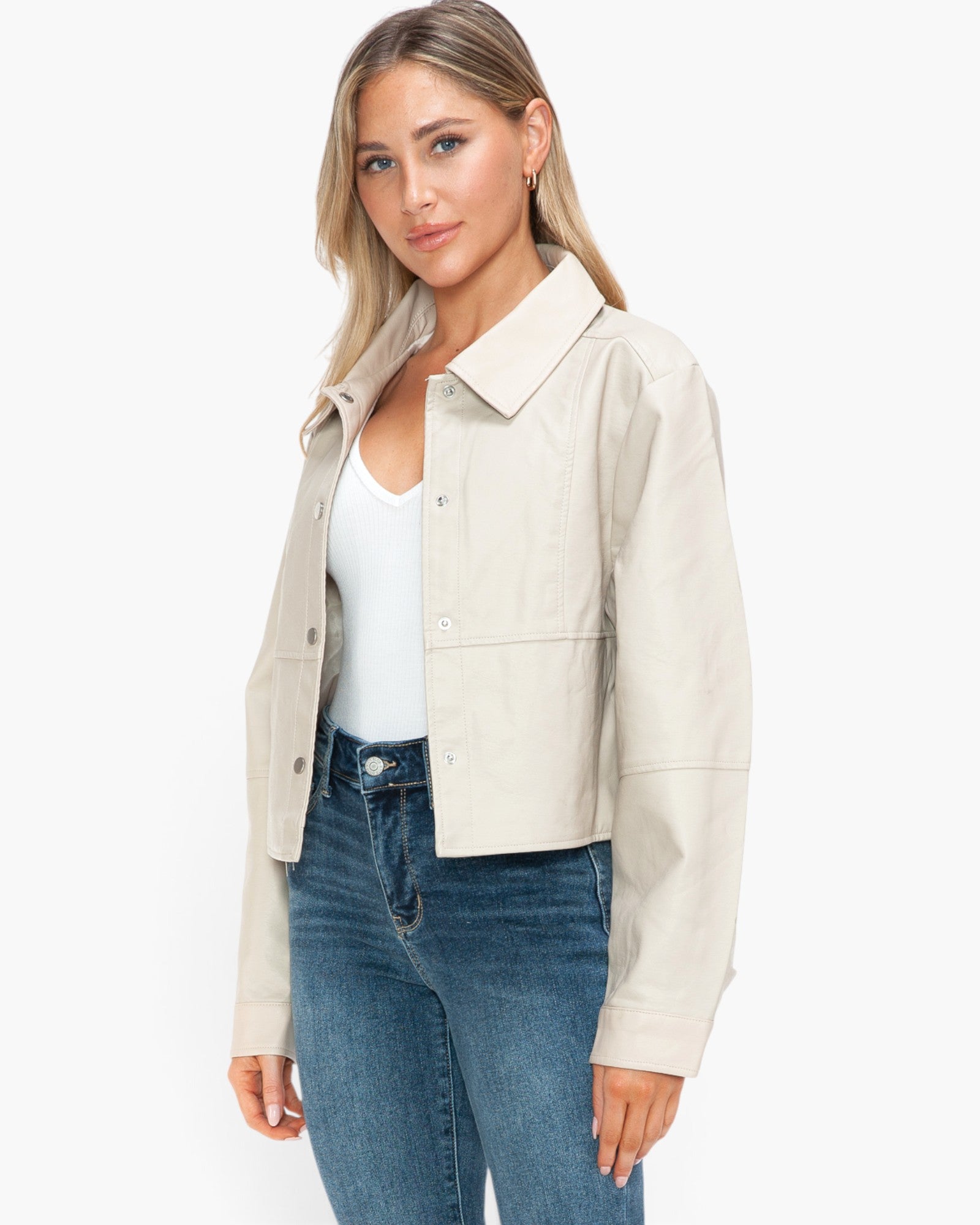 A woman modeling a light beige cropped utility jacket over a white tank top and blue jeans, photographed against a clean white studio background.