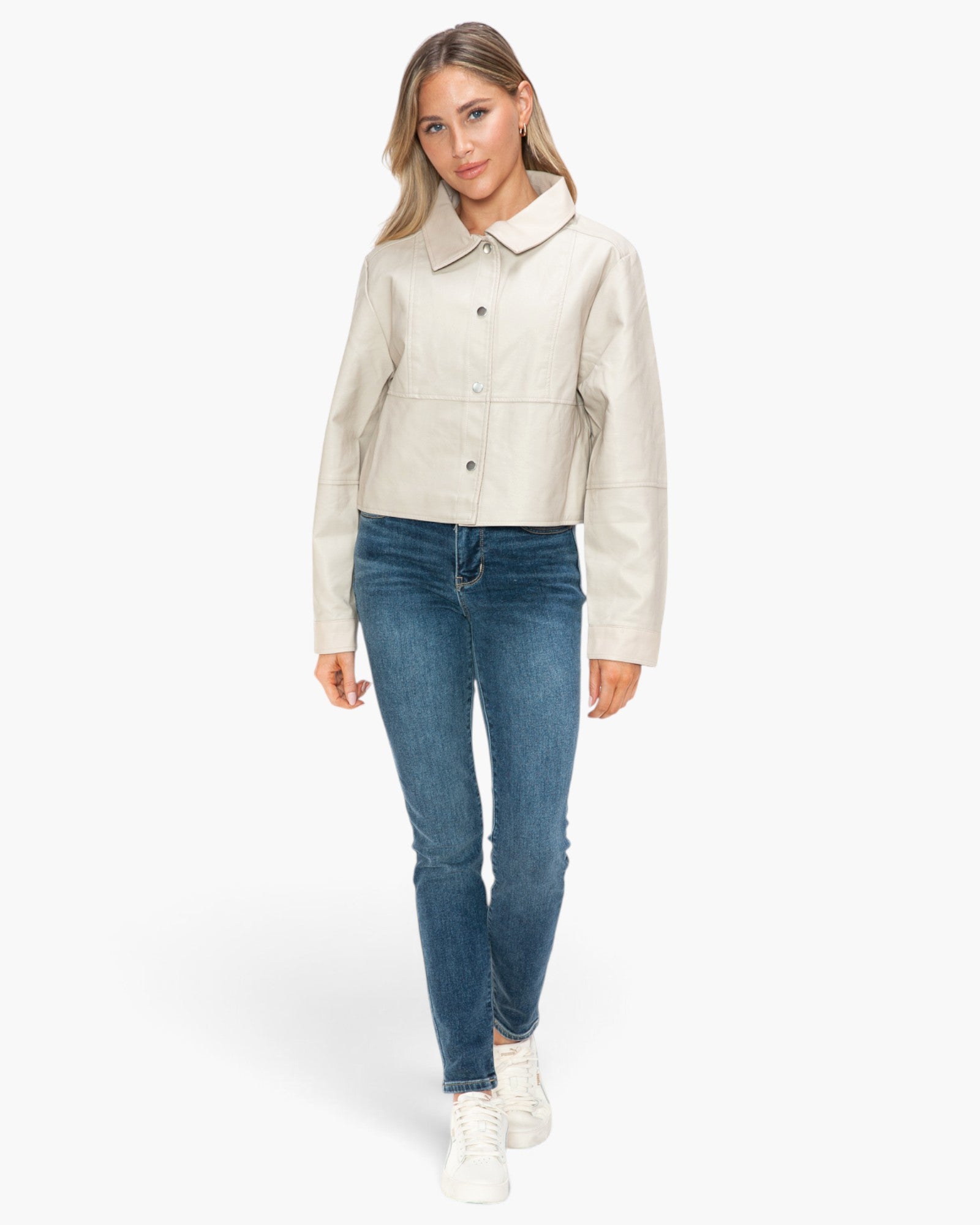 A woman modeling a cropped beige faux-leather jacket with a collar and button front, paired with blue skinny jeans and white sneakers, against a plain white background.