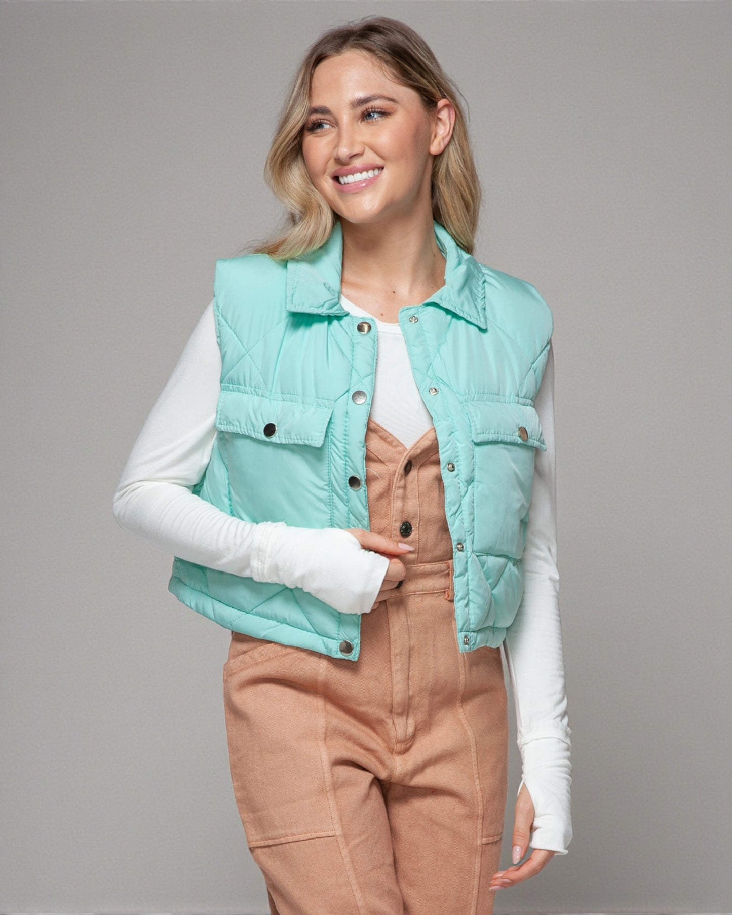 Woman wearing a light blue puffer vest over a white shirt and brown pants on a gray background
