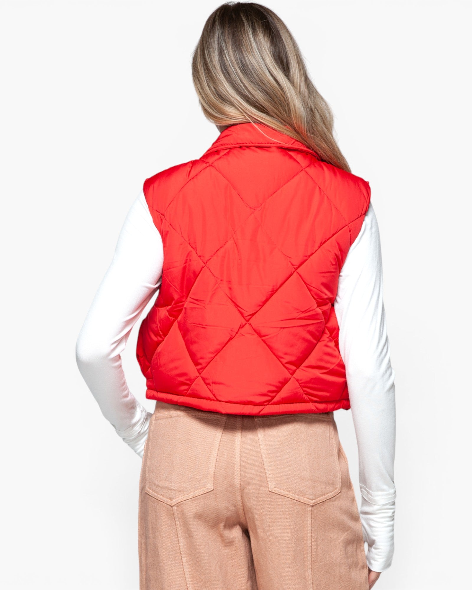 A woman shown from the back wearing a bright red quilted puffer vest over a white long-sleeve top and beige pants, against a plain light gray studio background.