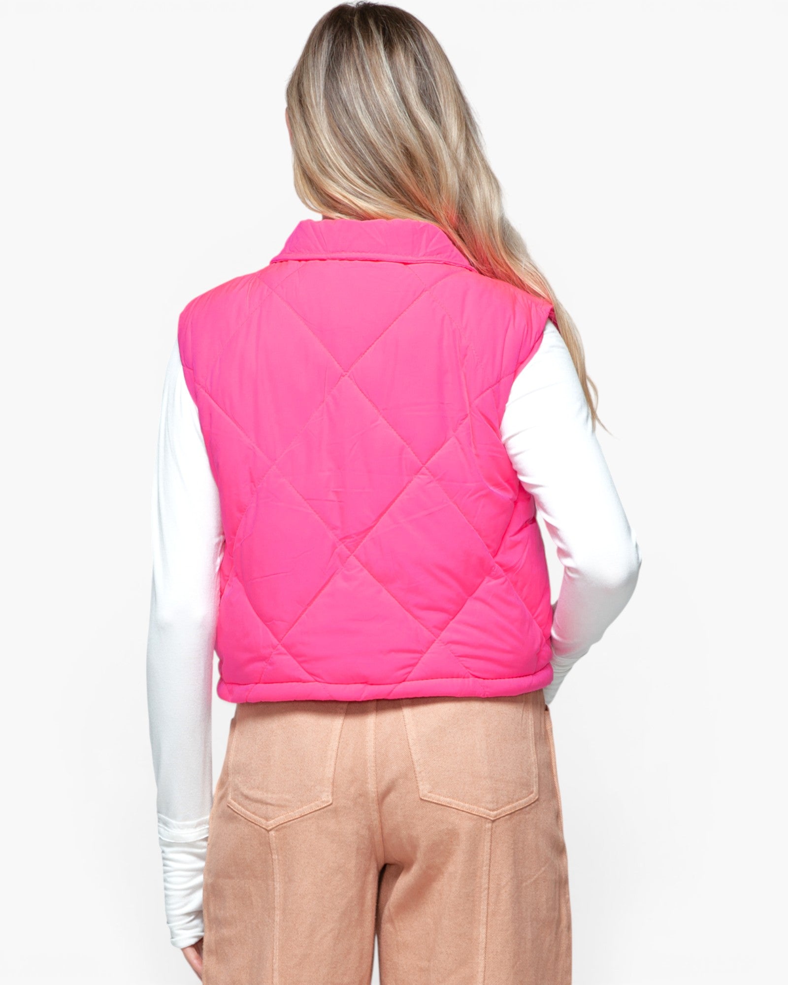 A woman viewed from the back wearing a bright pink quilted sleeveless vest over a white long-sleeve top and tan pants, against a plain light background.