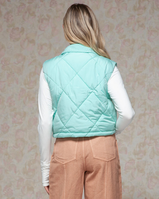 Person wearing a light blue quilted vest over a white shirt and brown pants against a floral-patterned wall.