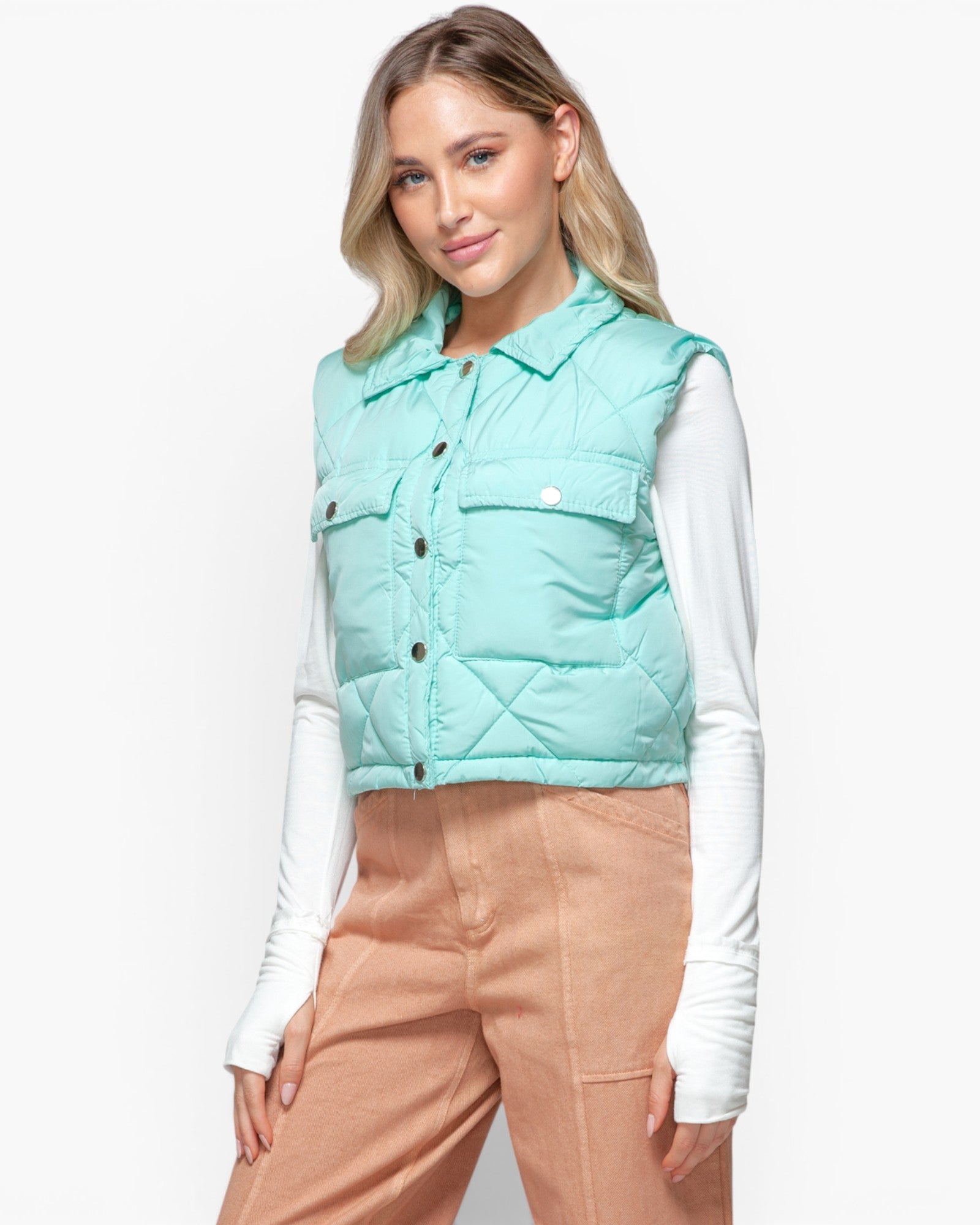 A woman wearing a mint green quilted puffer vest over a white long-sleeve shirt and tan pants, photographed against a clean white studio background.