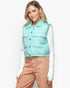 A woman wearing a mint green quilted puffer vest over a white long-sleeve shirt and tan pants, photographed against a clean white studio background.