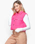 A woman wearing a bright pink sleeveless puffer vest over a white long-sleeve top and tan pants, posed against a light gray studio background.