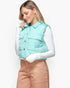 A woman wearing a mint green puffer vest over a white long-sleeve shirt and tan pants, photographed against a plain light gray studio background.
