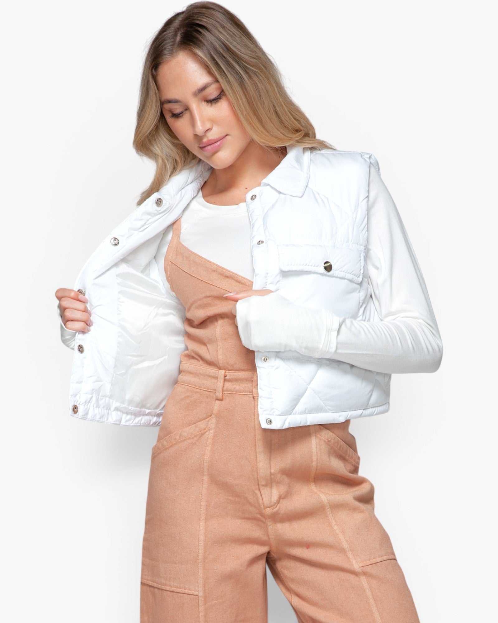 A woman wearing a white cropped puffer jacket over a white top and tan corduroy jumpsuit, posing against a light gray studio background.