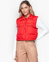 A woman wearing a bright red quilted puffer vest over a white long-sleeve top and tan pants, photographed against a clean white studio background.