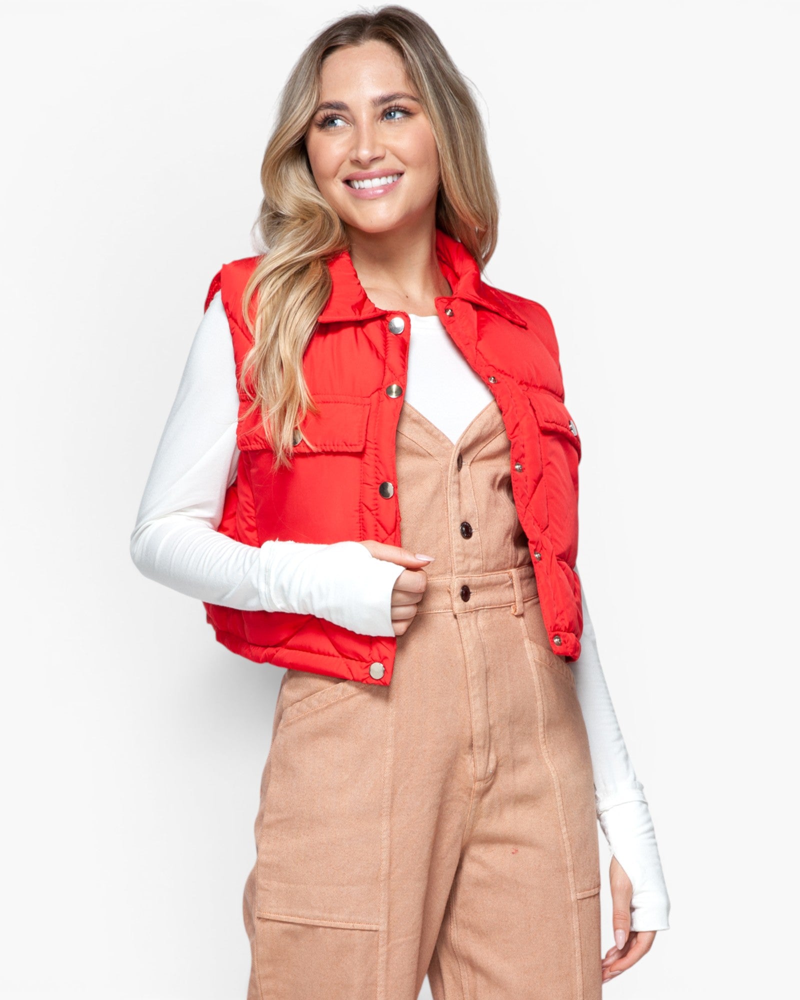 A woman wearing a bright red sleeveless puffer vest over a white long-sleeve top and tan corduroy overalls, photographed against a clean light gray studio background.