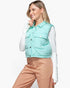 A woman wearing a mint green quilted puffer vest over a white long-sleeve top and peach pants, posing against a solid white studio background.