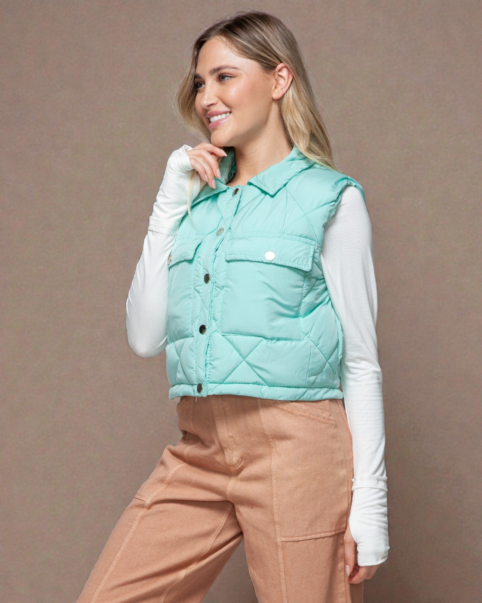 Woman wearing a light blue puffer vest over a white shirt with beige pants on a brown background