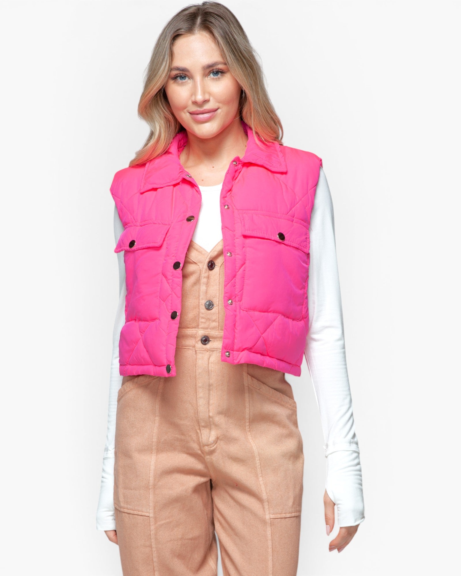 A woman modeling a bright pink puffer vest over a white long-sleeve top and tan corduroy pants, photographed against a light gray studio background.