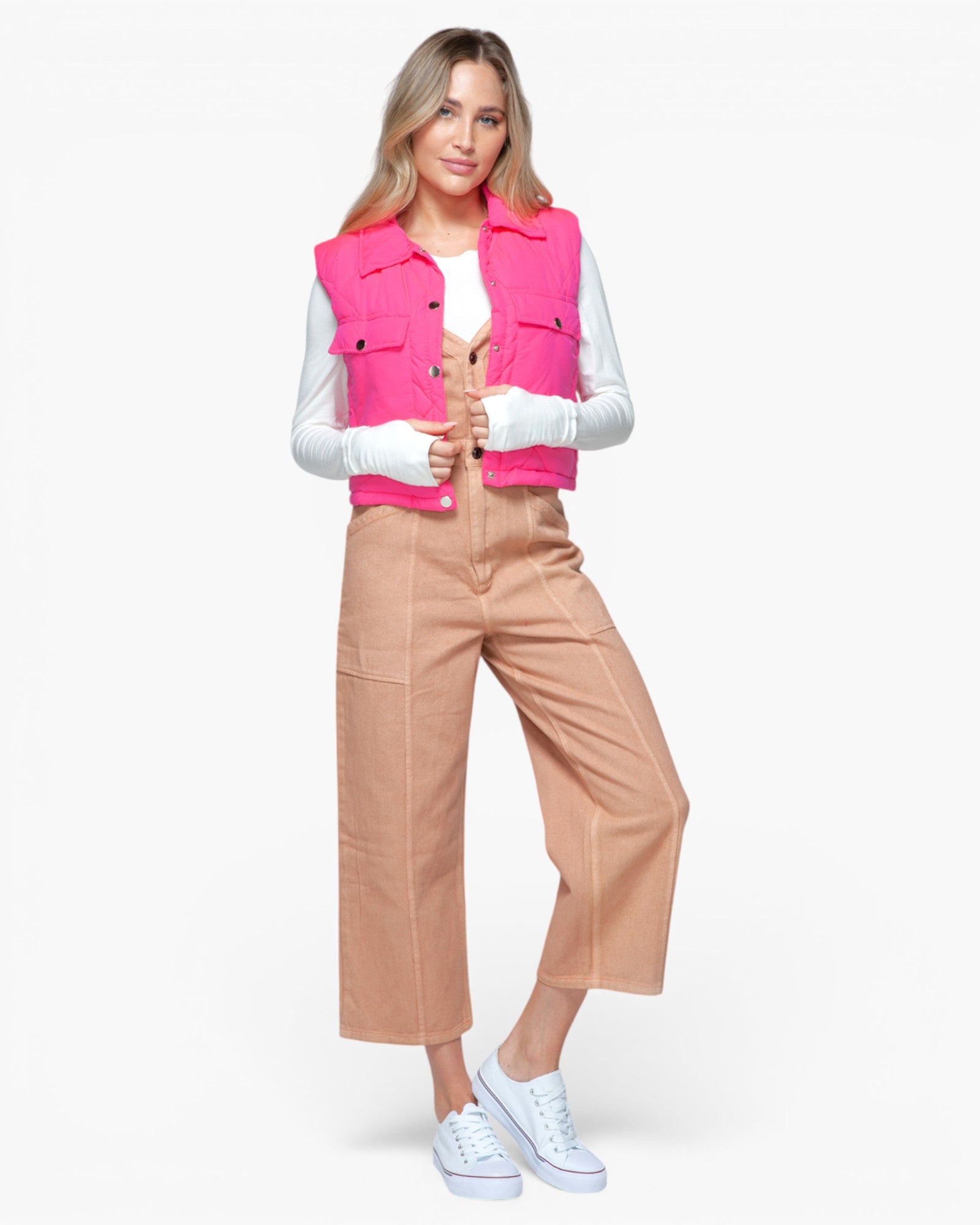 A woman modeling a bright pink sleeveless vest over a white long-sleeve top, paired with tan cropped pants and white sneakers, against a light gray studio background.