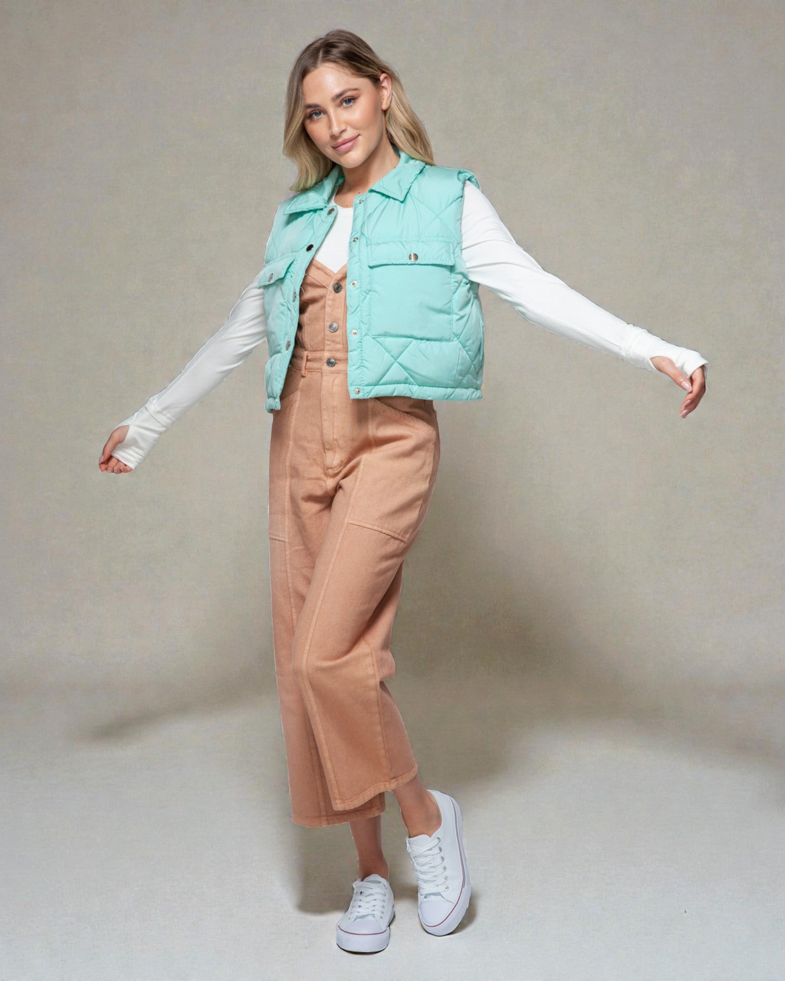 Woman wearing a light blue puffer vest over a white shirt and beige pants on a plain background