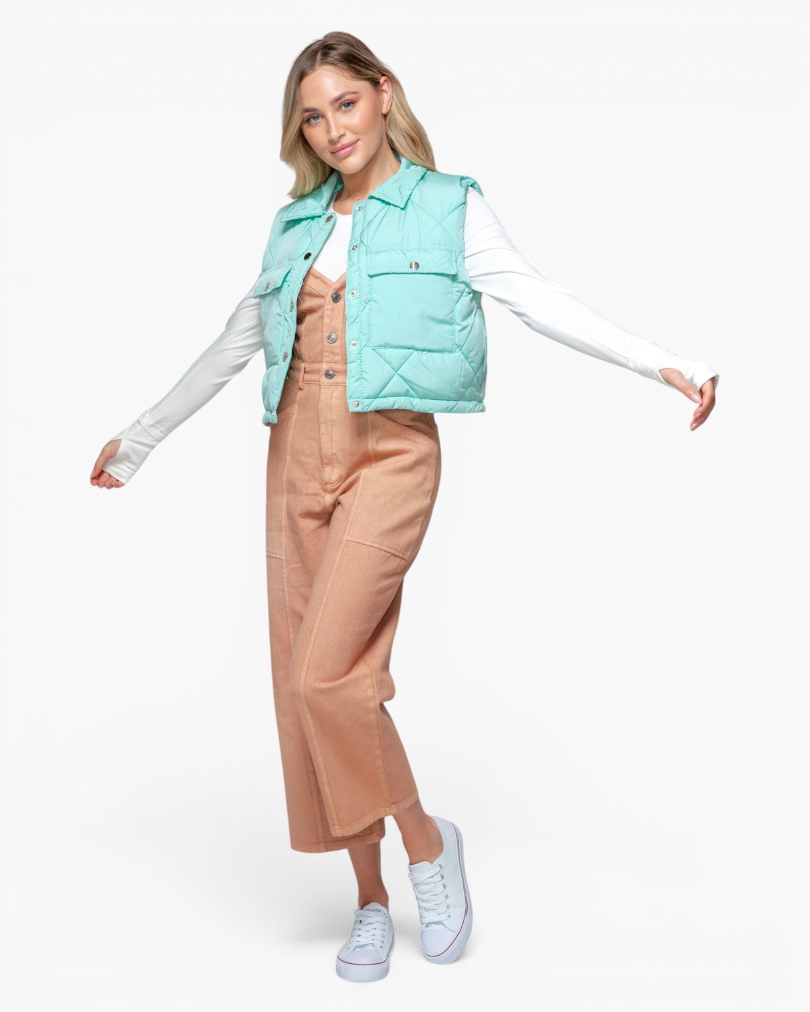 A woman modeling a mint green puffer vest over a white long-sleeve shirt and tan jumpsuit, paired with white sneakers, against a plain white studio background.