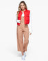 A woman modeling a bright red sleeveless puffer vest over a white long-sleeve top and tan wide-leg pants, paired with white sneakers, on a light gray studio background.