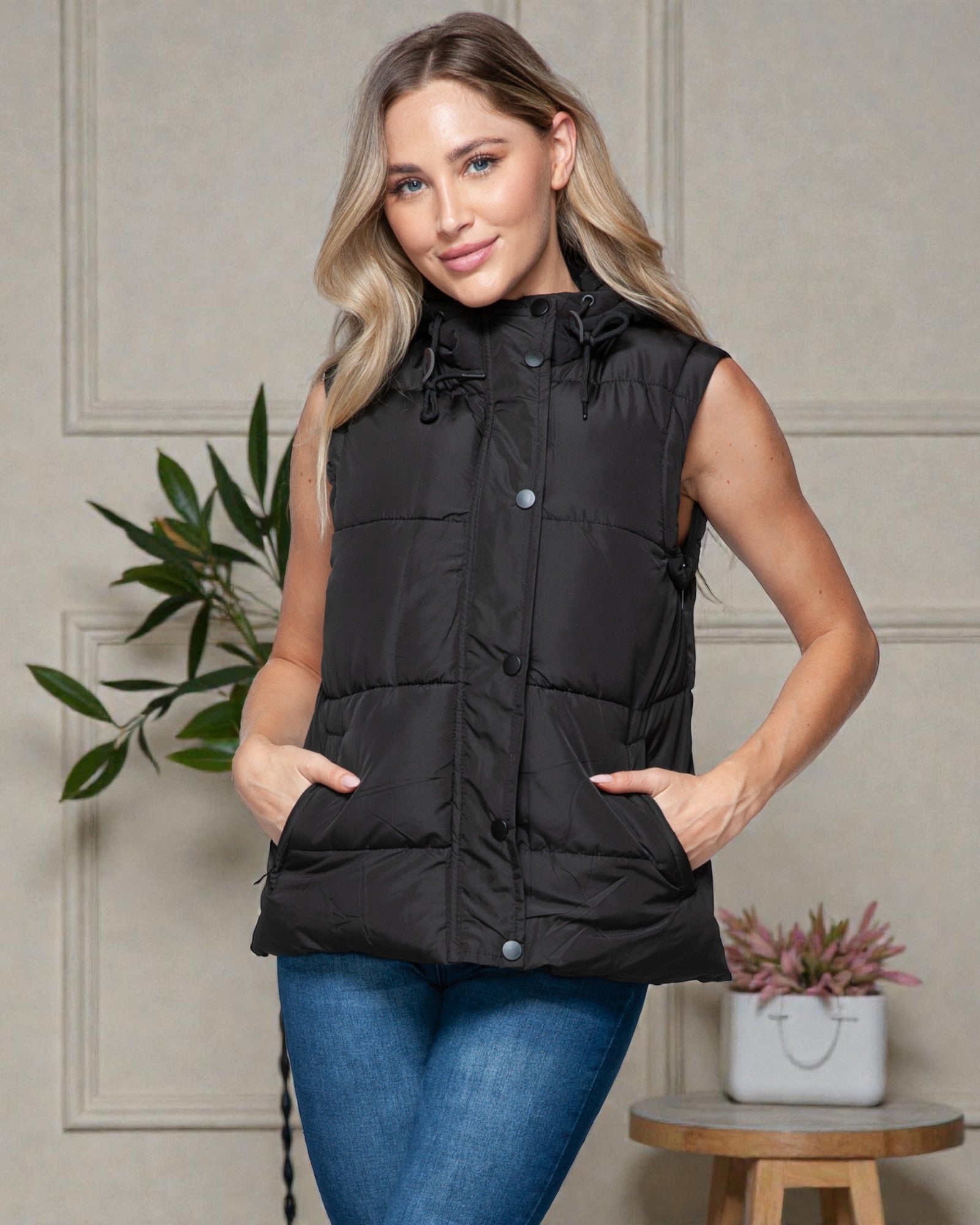 Woman wearing a black puffer vest against a neutral wall with plants.