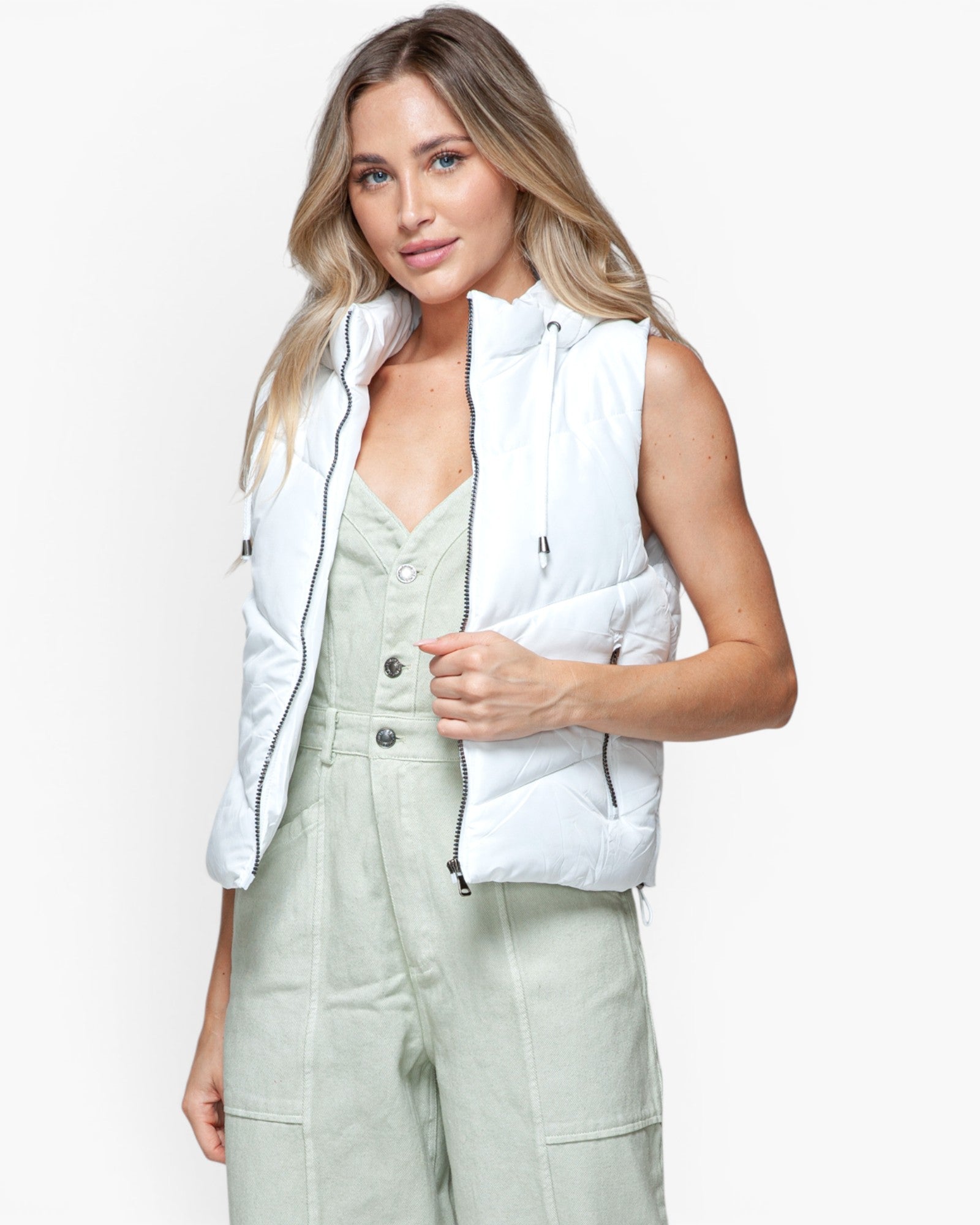 A woman modeling a sleeveless white puffer vest over a sage green jumpsuit, photographed against a clean white studio background.