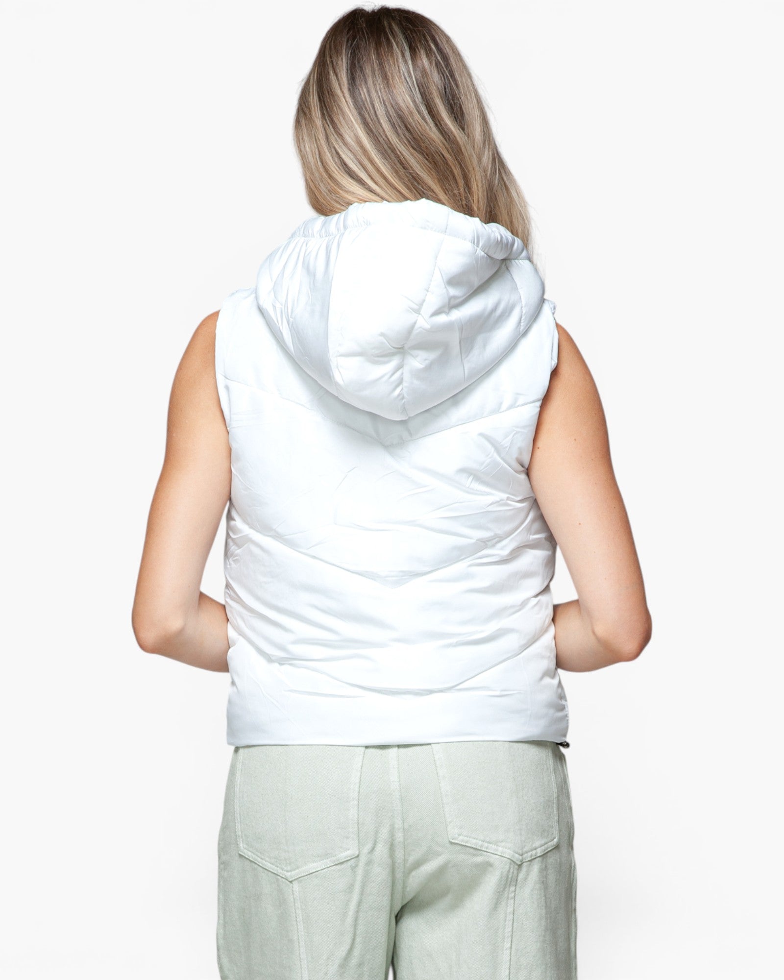 A woman viewed from behind wearing a white sleeveless puffer vest with a hood and light sage green pants, against a plain white studio background.
